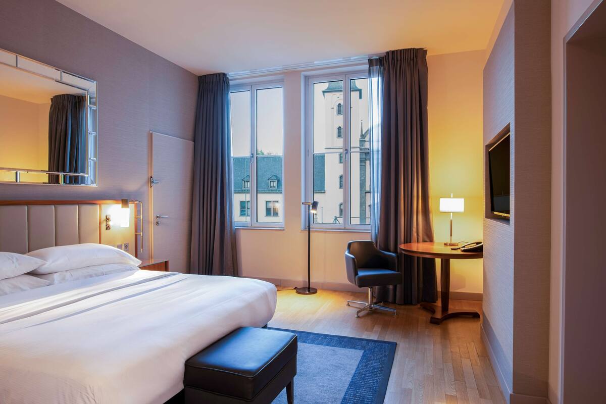 Habitación de hotel con una cama grande, un escritorio con una silla, una lámpara de escritorio y una ventana con cortinas.