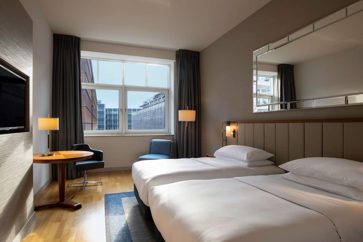 Habitación de hotel con dos camas, un escritorio, una silla, una ventana con cortinas y un espejo grande en la pared.