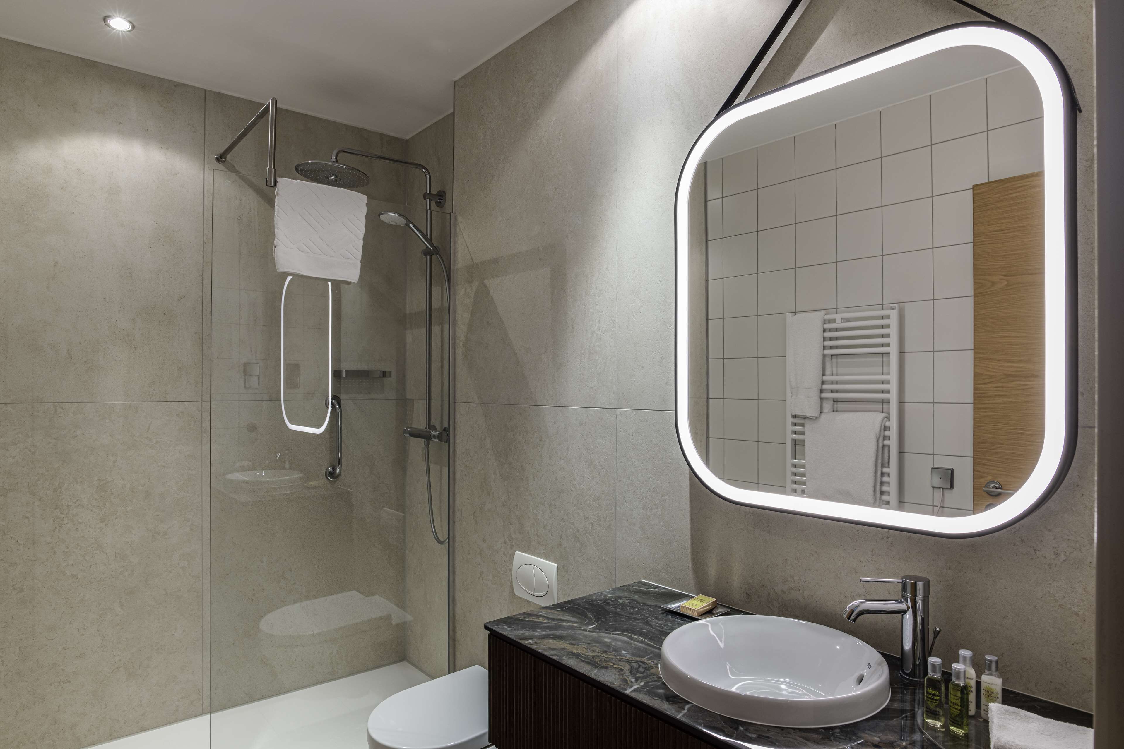 A modern bathroom with a glass-enclosed shower, a white toilet, a marble sink, and a mirror with a lighted frame.