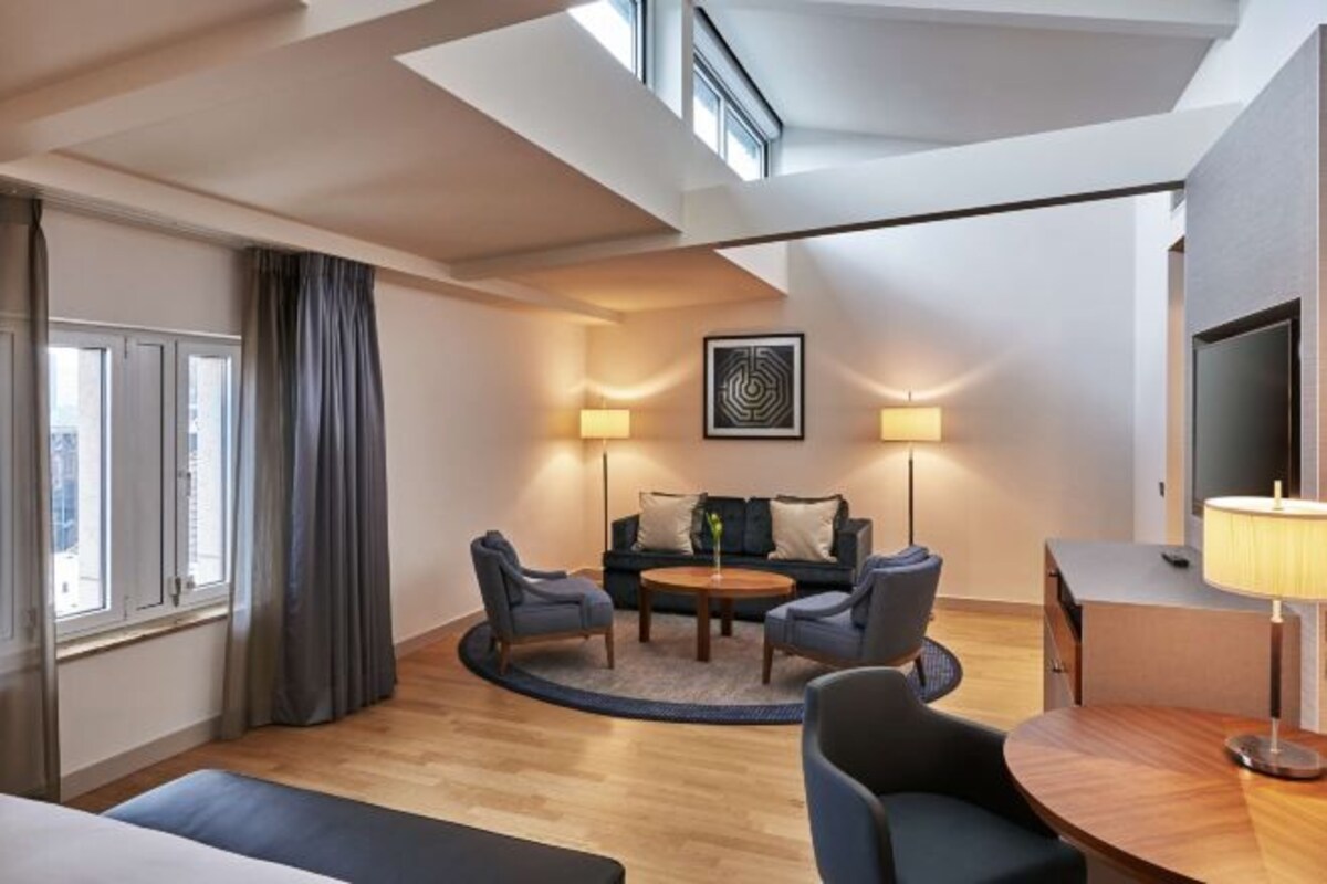 A modern living room with a round coffee table, two armchairs, a sofa, and a TV. There is a window with curtains, a ceiling with skylights, and a lamp on the side table.