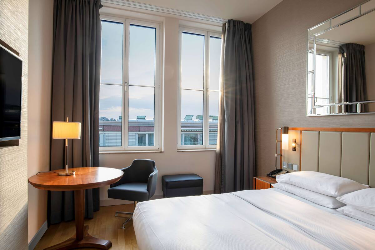 Habitación de hotel con una cama, un escritorio con lámpara, una silla, una mesita y un ventanal grande con cortinas.