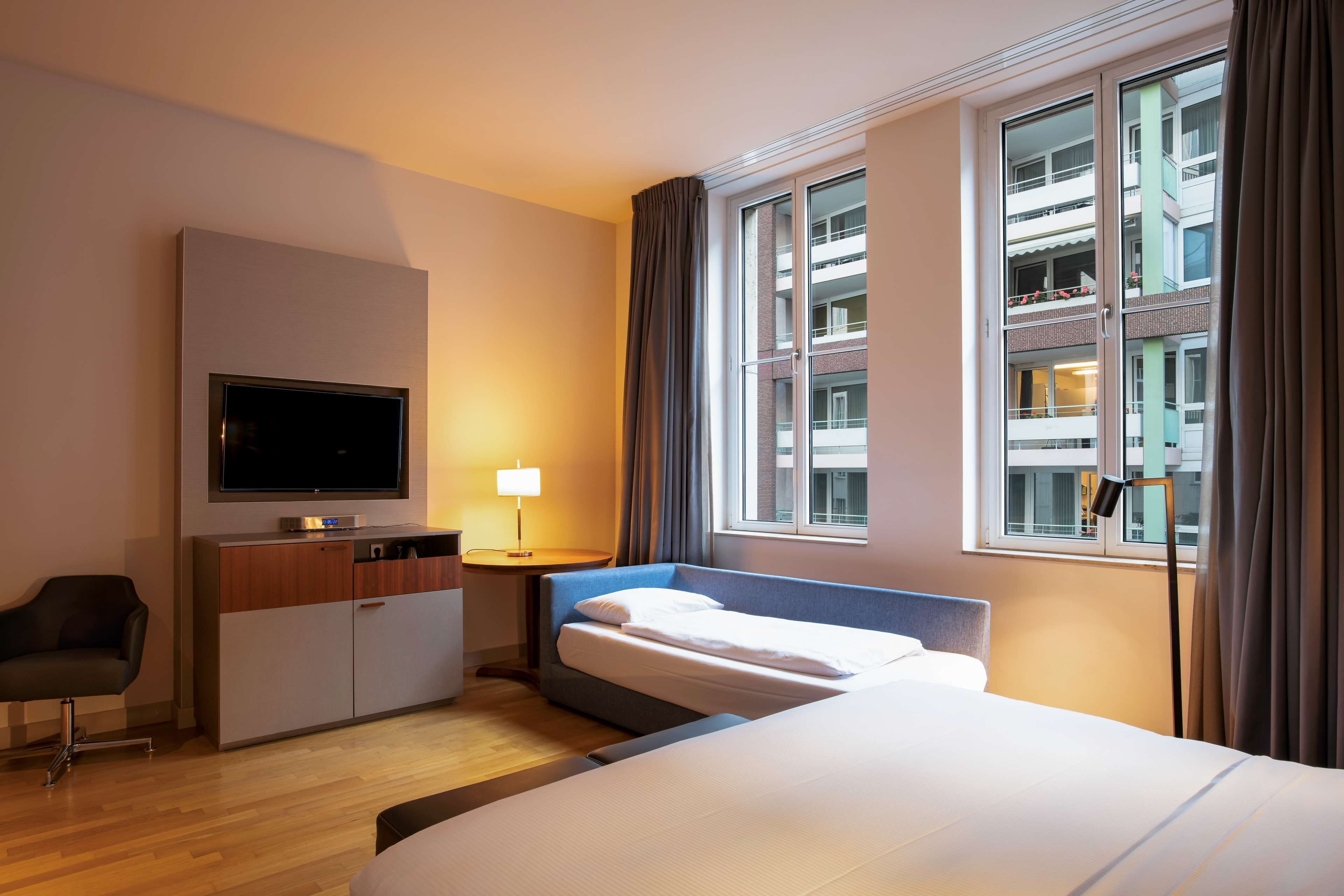 A hotel room with a bed, a desk with a TV, a chair, a lamp, and a window with curtains.