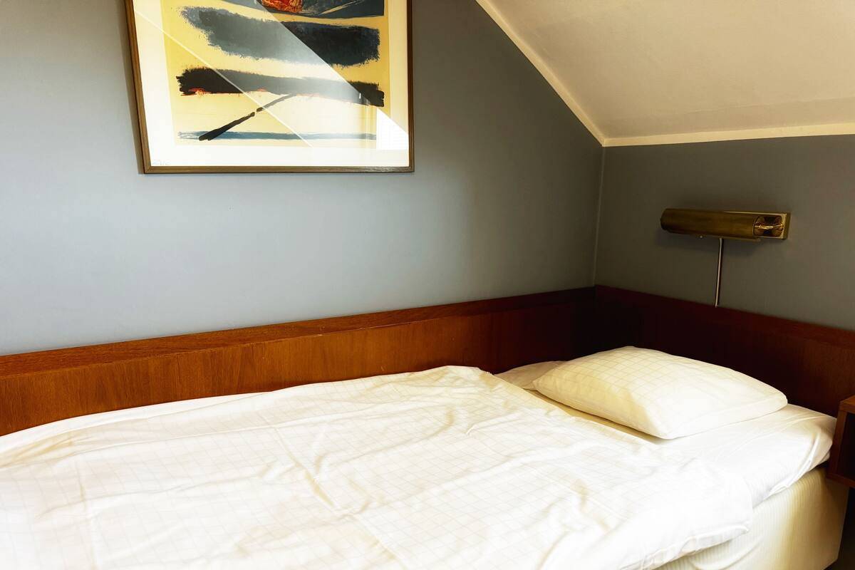A neatly made bed with a wooden headboard, a bedside table with a lamp, and a framed abstract painting on the wall.
