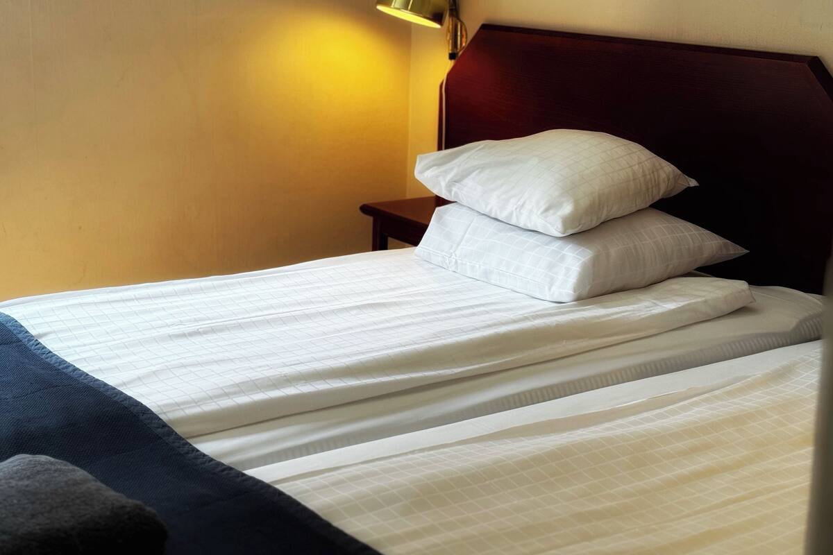 A neatly made bed with white linens and two pillows, a wooden headboard, and a bedside lamp.