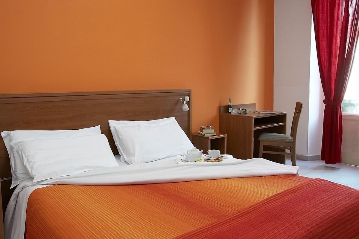 A hotel room with a bed, a nightstand, a chair, and a window with red curtains.