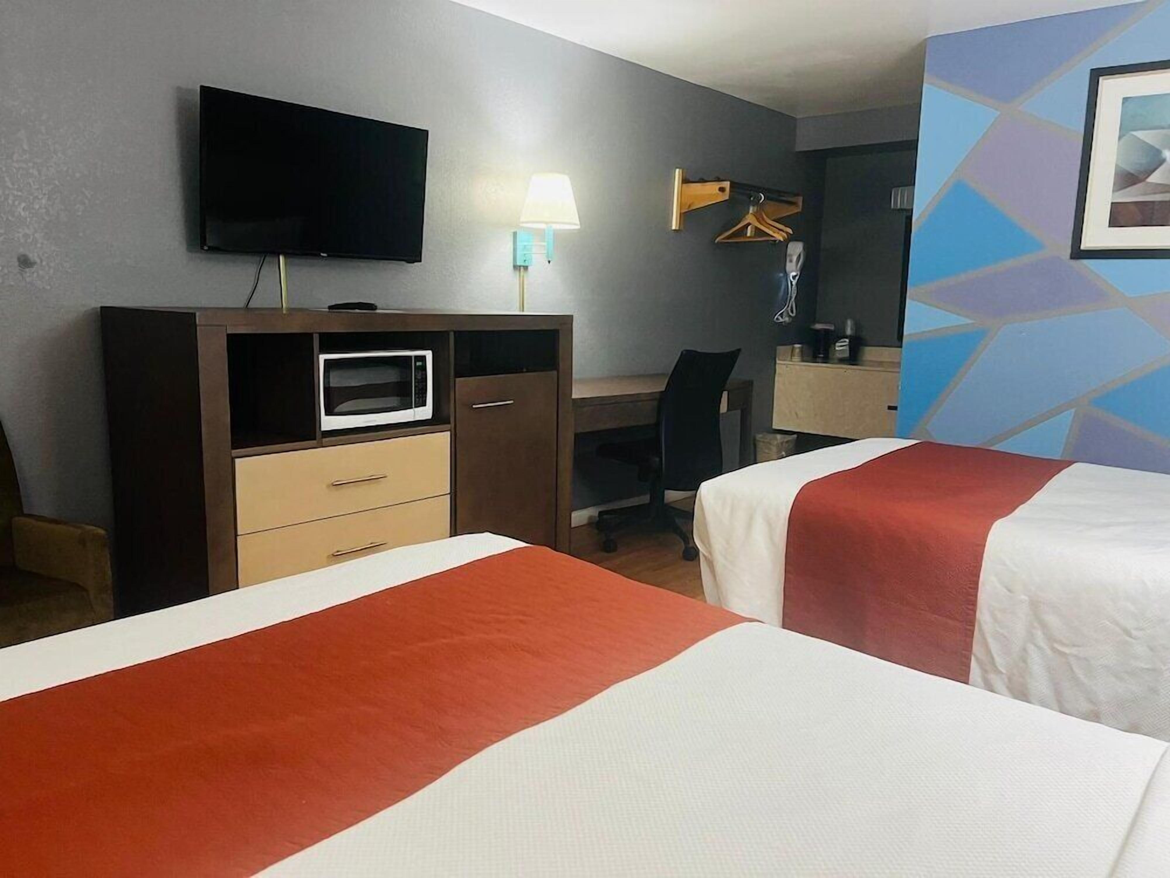 A hotel room with two beds, a desk, a chair, a TV, a microwave, and a wall-mounted lamp.