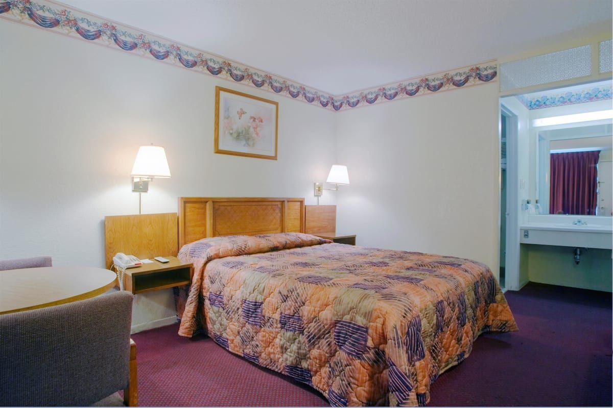 A hotel room with a bed, two bedside lamps, a wooden headboard, a desk with a chair, a telephone, and a painting on the wall.