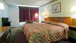 A hotel room with a large bed, a desk with a lamp, a television, and a door with purple curtains.