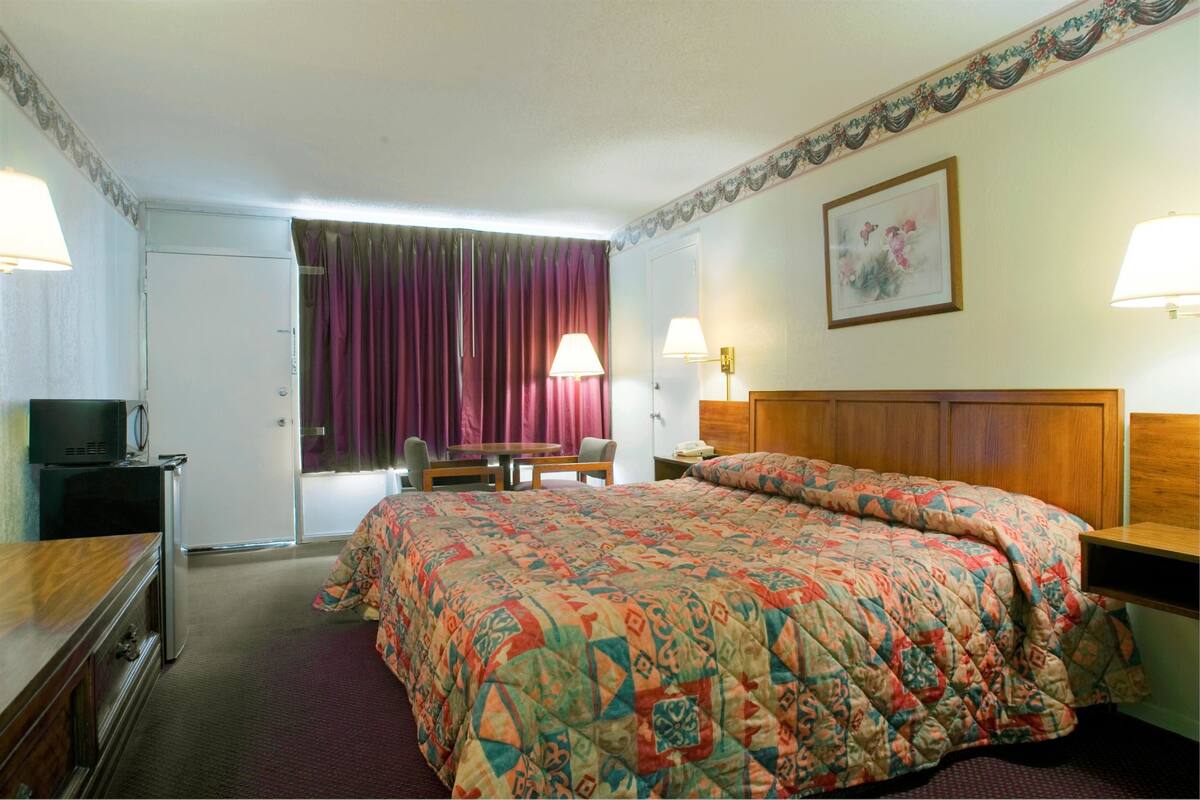 A hotel room with a large bed, a desk with a lamp, a television, and a door with purple curtains.