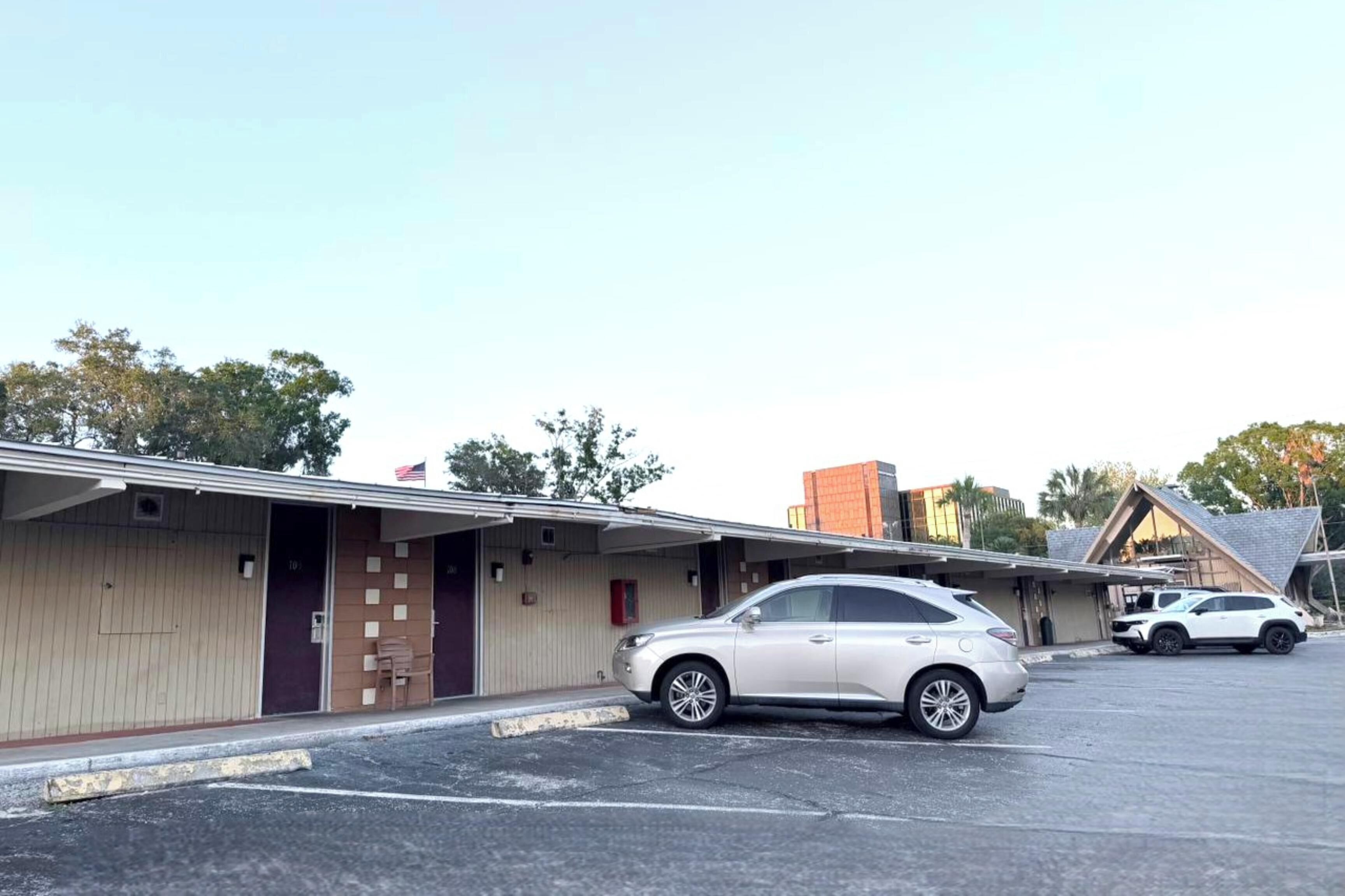 Un motel con una edificación de un piso, un área de estacionamiento techada y una fila de habitaciones con puertas.