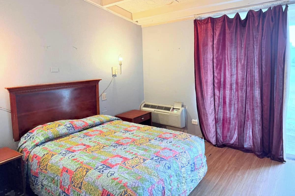 A bedroom with a wooden headboard, a colorful quilt, a bedside table, an air conditioning unit, and a window with a curtain.