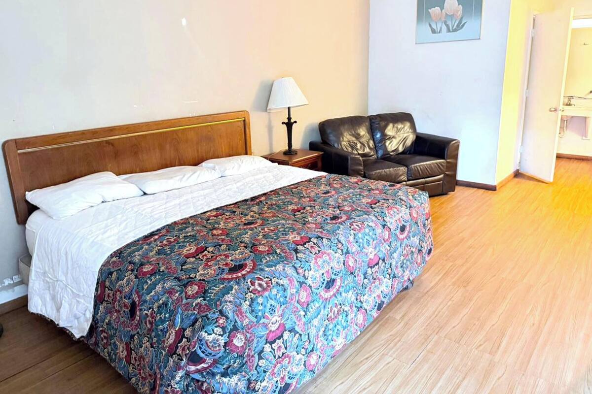 A bedroom with a bed, a nightstand, a lamp, a leather chair, and a painting on the wall.