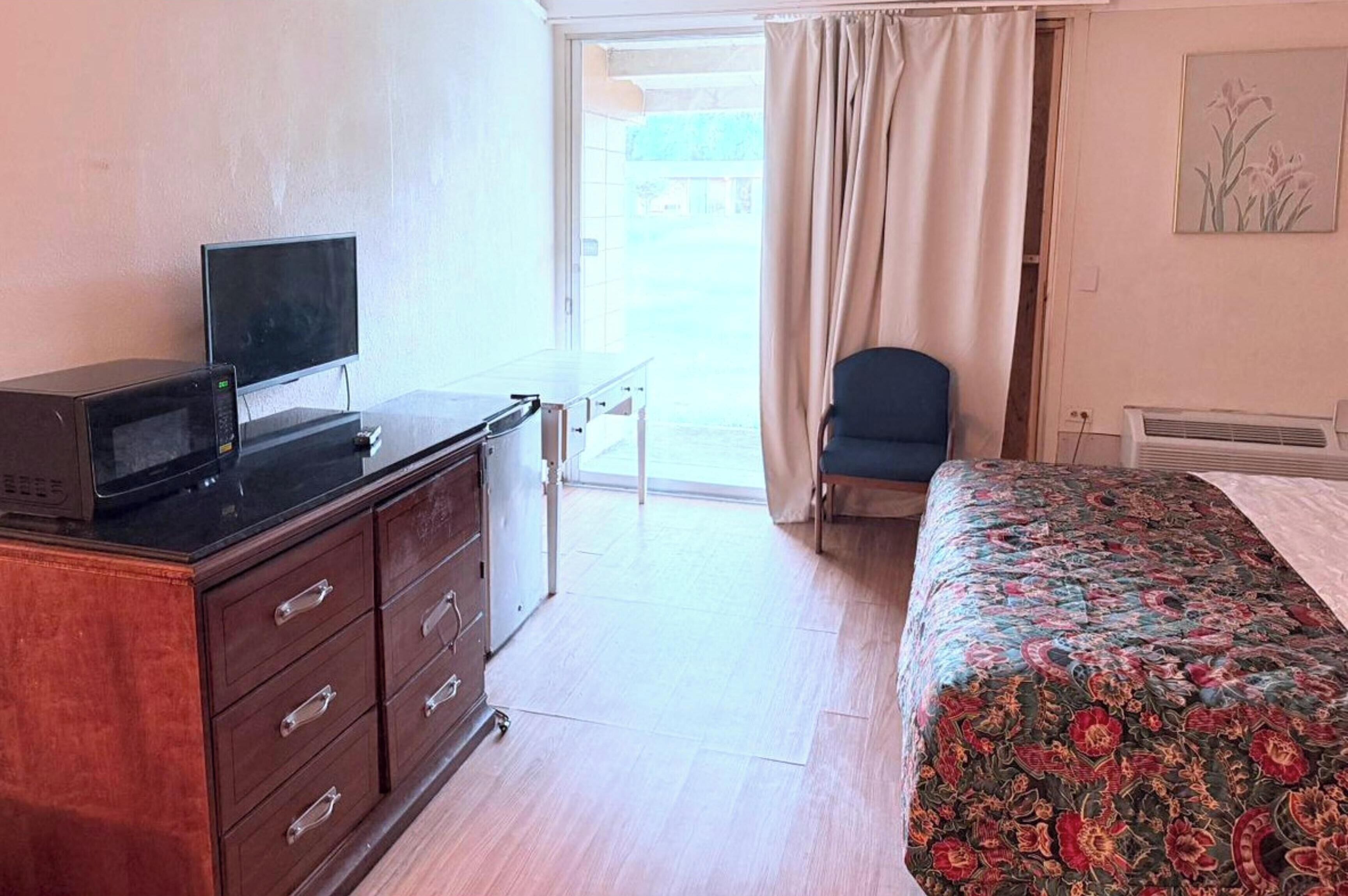 Et hotelværelse med en seng, et skrivebord med computer, en stol, et vindue med gardiner og en fastgjort aircondition.