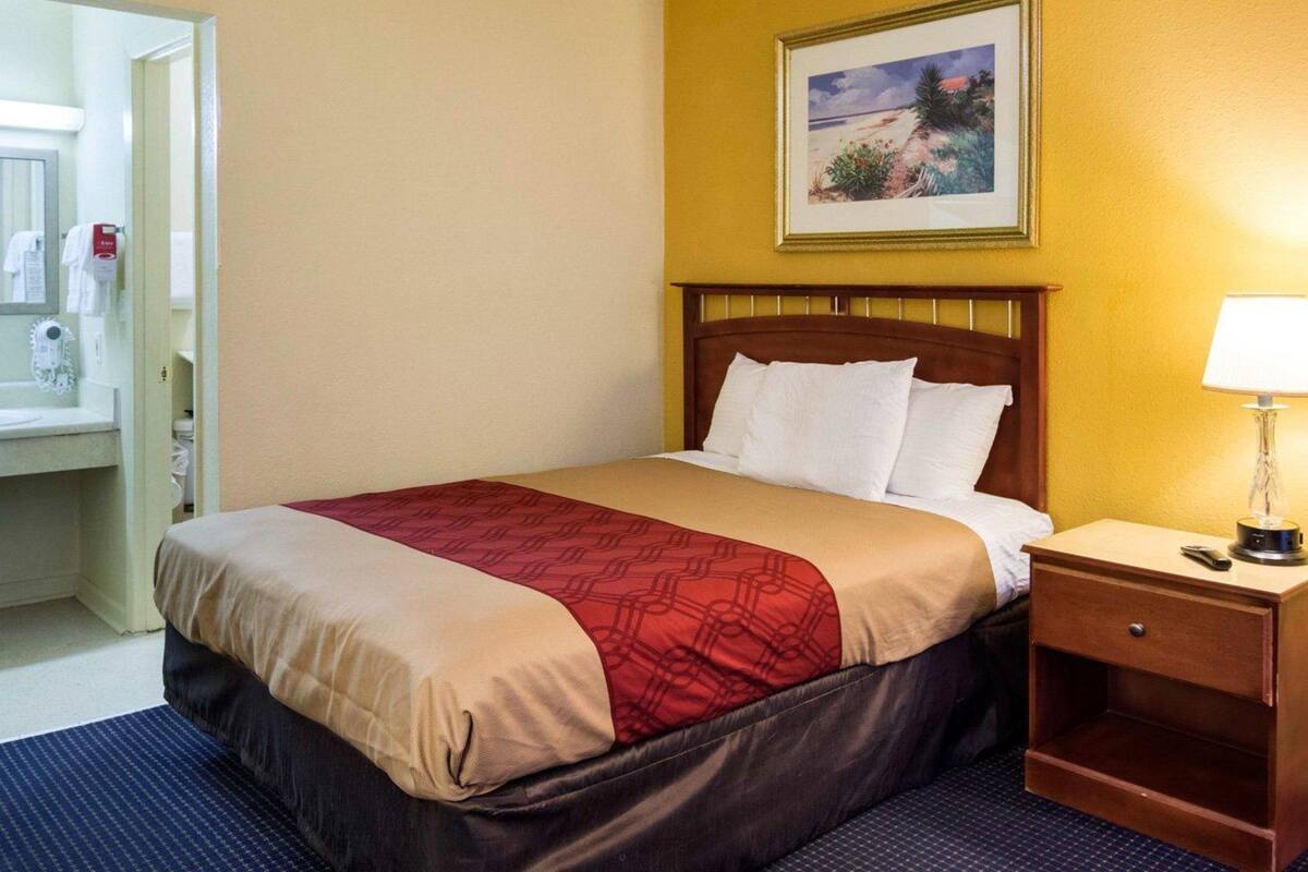 Una camera d'albergo con un letto, un comodino, una lampada, un quadro appeso e un bagno visibile attraverso una porta aperta.