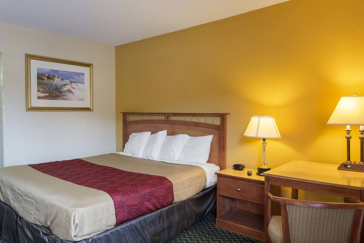 Una camera d'albergo con un letto, un comodino, una lampada, una sedia e un quadro appeso al muro.