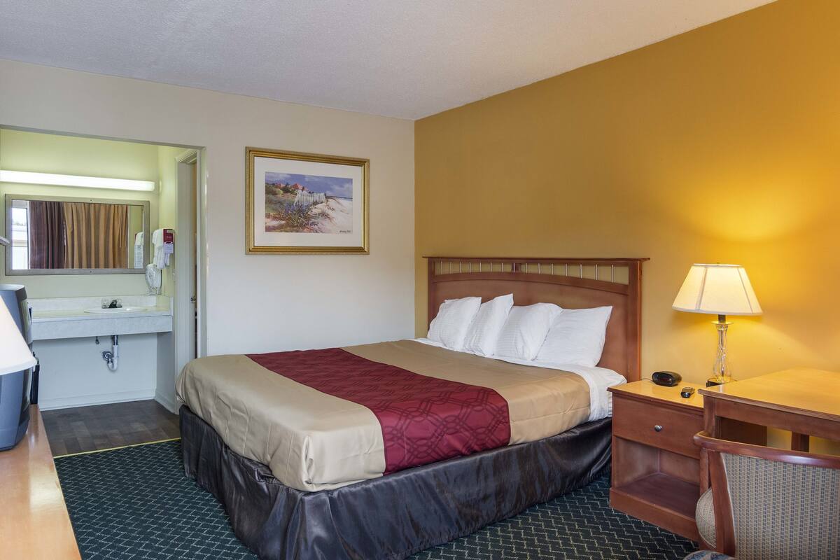 Una camera d'albergo con un letto, comodino, lampada e bagno con lavandino e specchio.