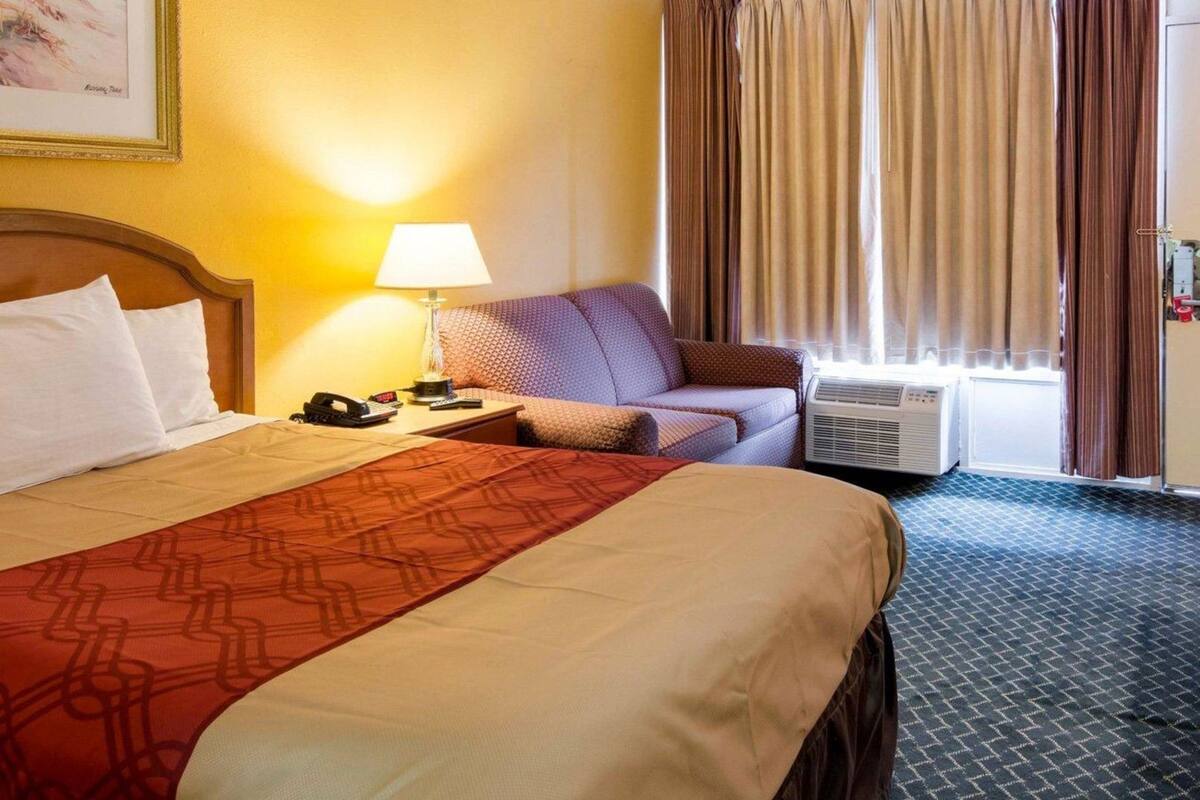 Una camera d'albergo con un letto, un divano, una lampada, un condizionatore d'aria e una finestra con tende.