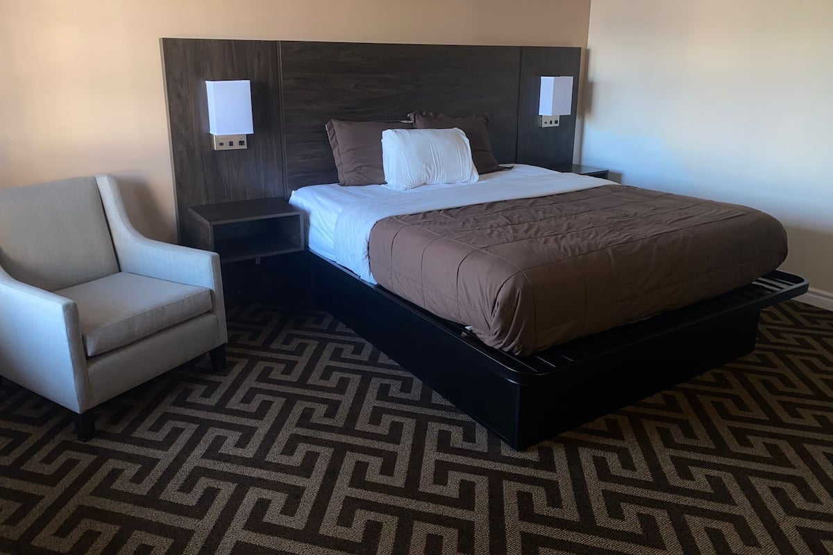A hotel room with a bed, a chair, a nightstand, and a patterned carpet.
