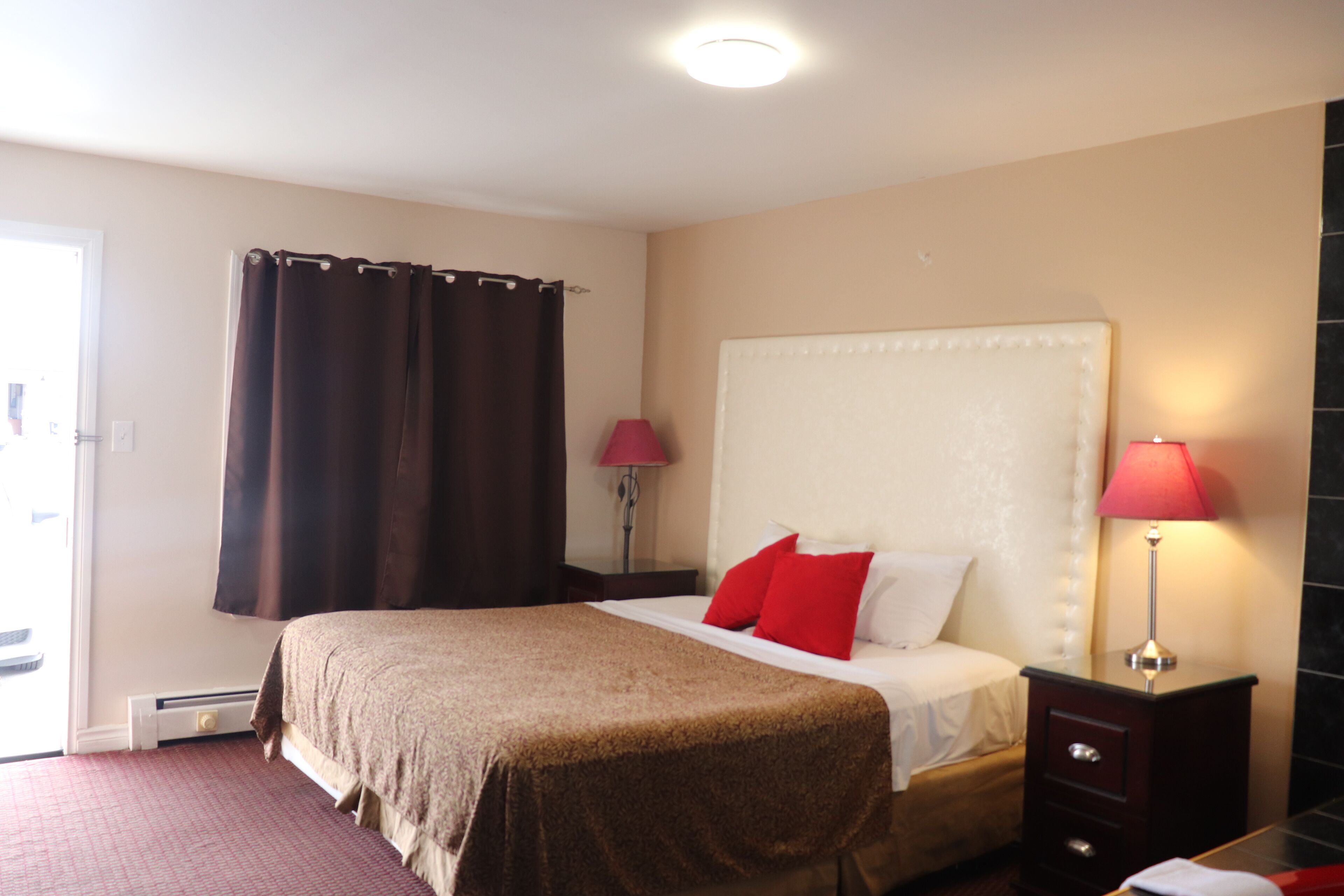 A hotel room with a large bed, a nightstand with a lamp, a window with curtains, and a door.