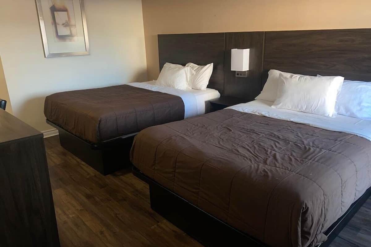 A hotel room with two beds, a desk, a nightstand, a wall-mounted lamp, and a framed picture.