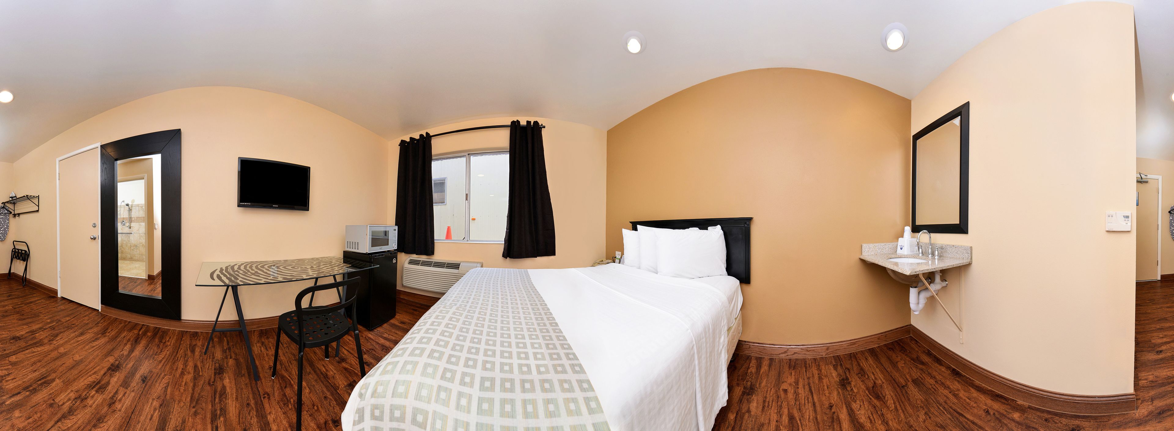 Habitación de hotel con cama, escritorio con silla, televisión, baño con lavamanos y espejo.