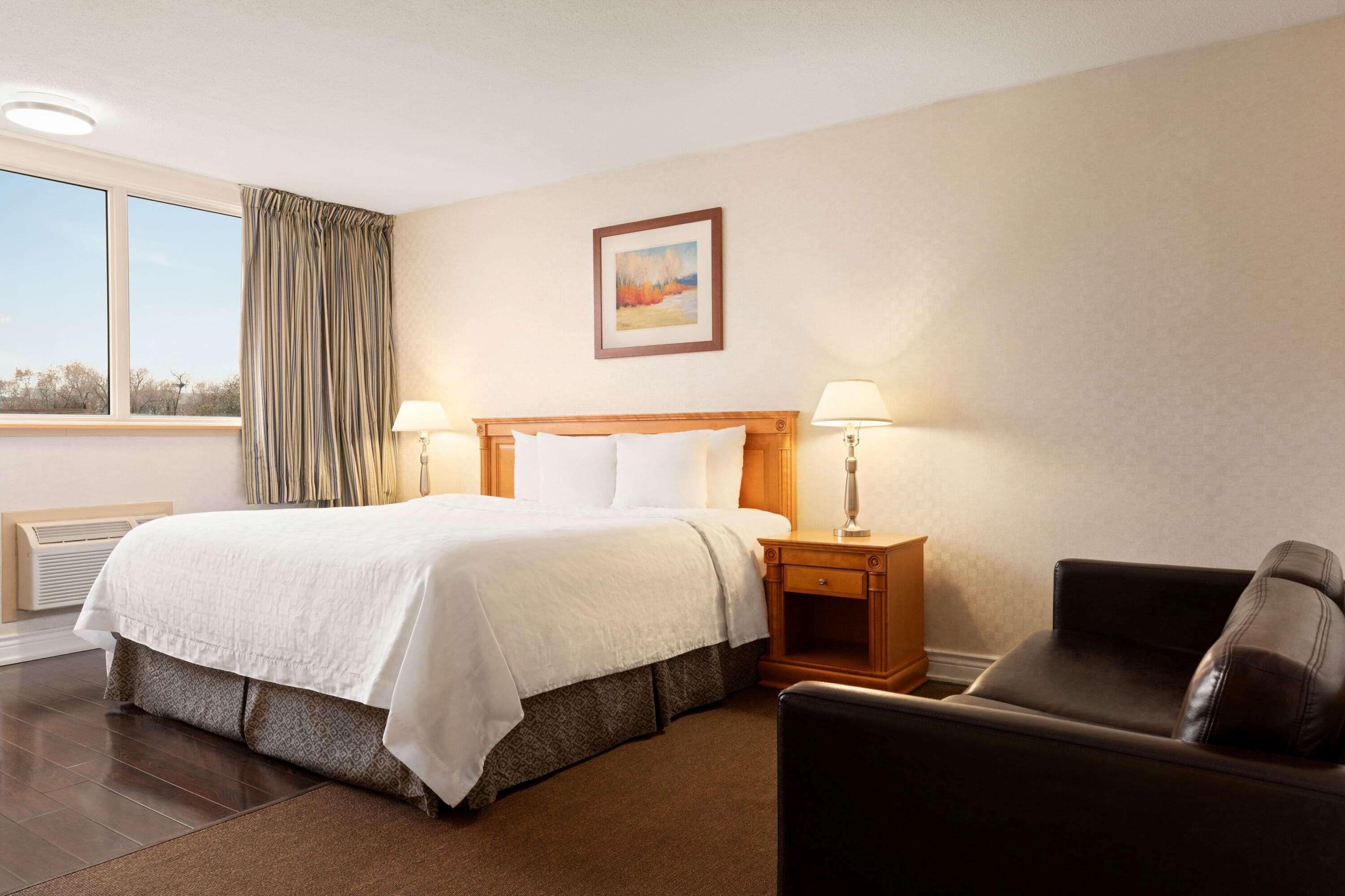 A hotel room with a large bed, two bedside lamps, a chair, a window with curtains, and a painting on the wall.