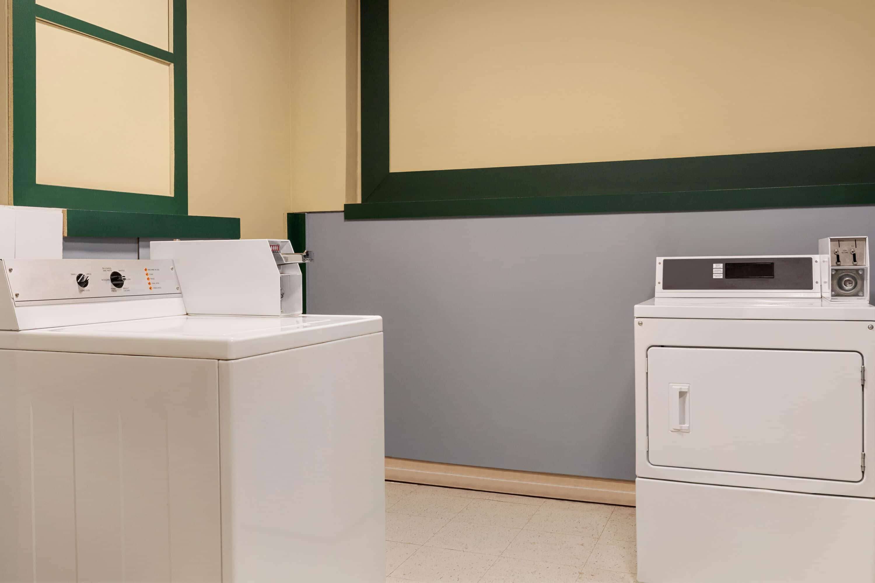 A laundry room with two white washing machines and a dryer, a control panel, and a wall-mounted shelf.