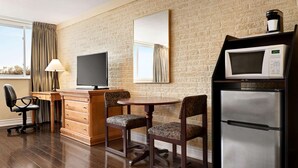 A hotel room with a brick wall, a flat-screen TV, a microwave, a refrigerator, a wooden desk, two upholstered chairs, a lamp, and a window with curtains.