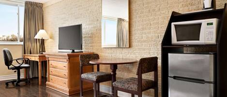 Habitación de hotel con pared de ladrillo, televisión de pantalla plana, microondas, refrigerador, escritorio de madera, dos sillones tapizados, lámpara y ventana con cortinas.