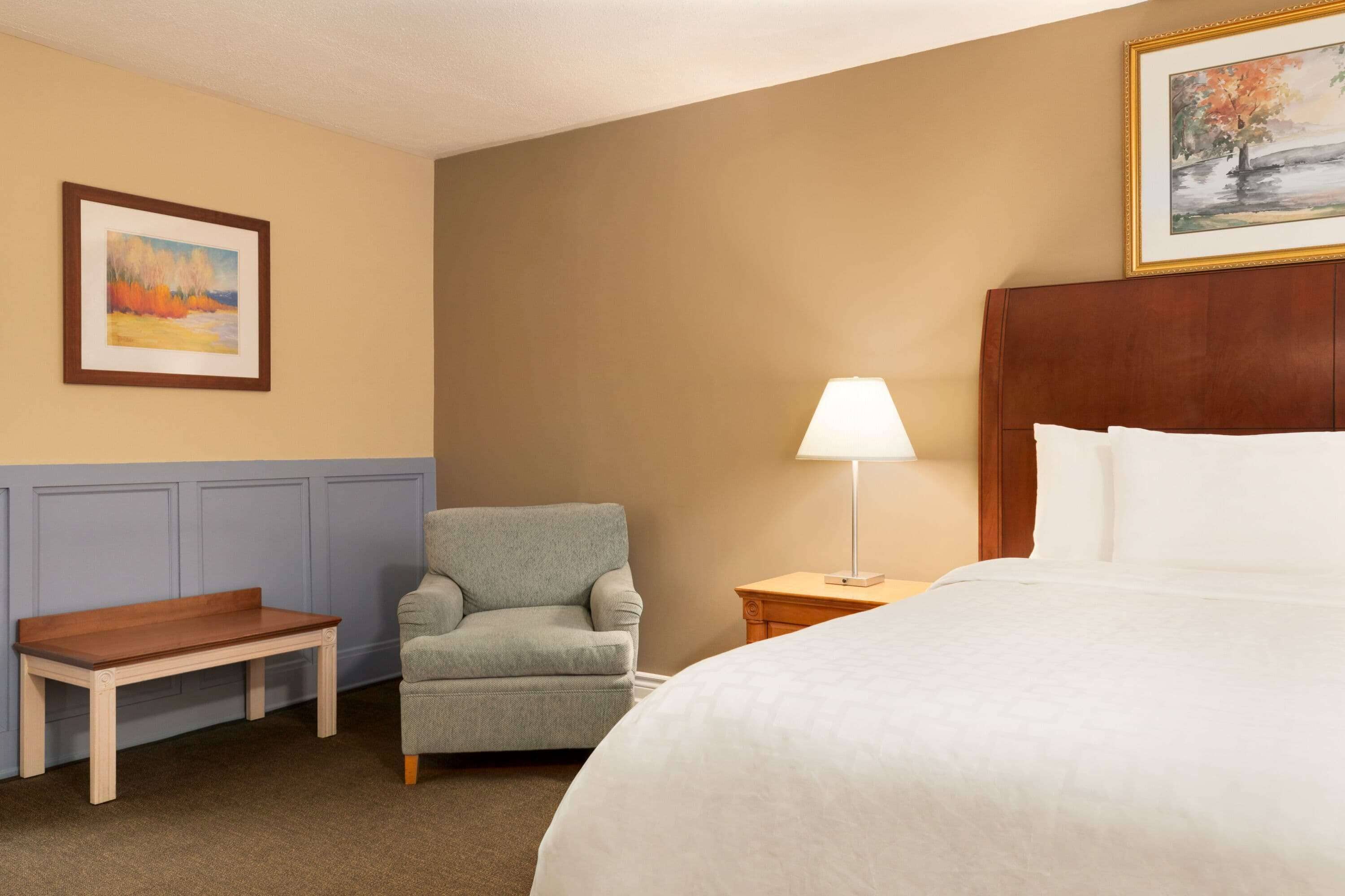 Une chambre d’hôtel avec un lit, une chaise, une petite table, une lampe et deux tableaux encadrés accrochés aux murs.