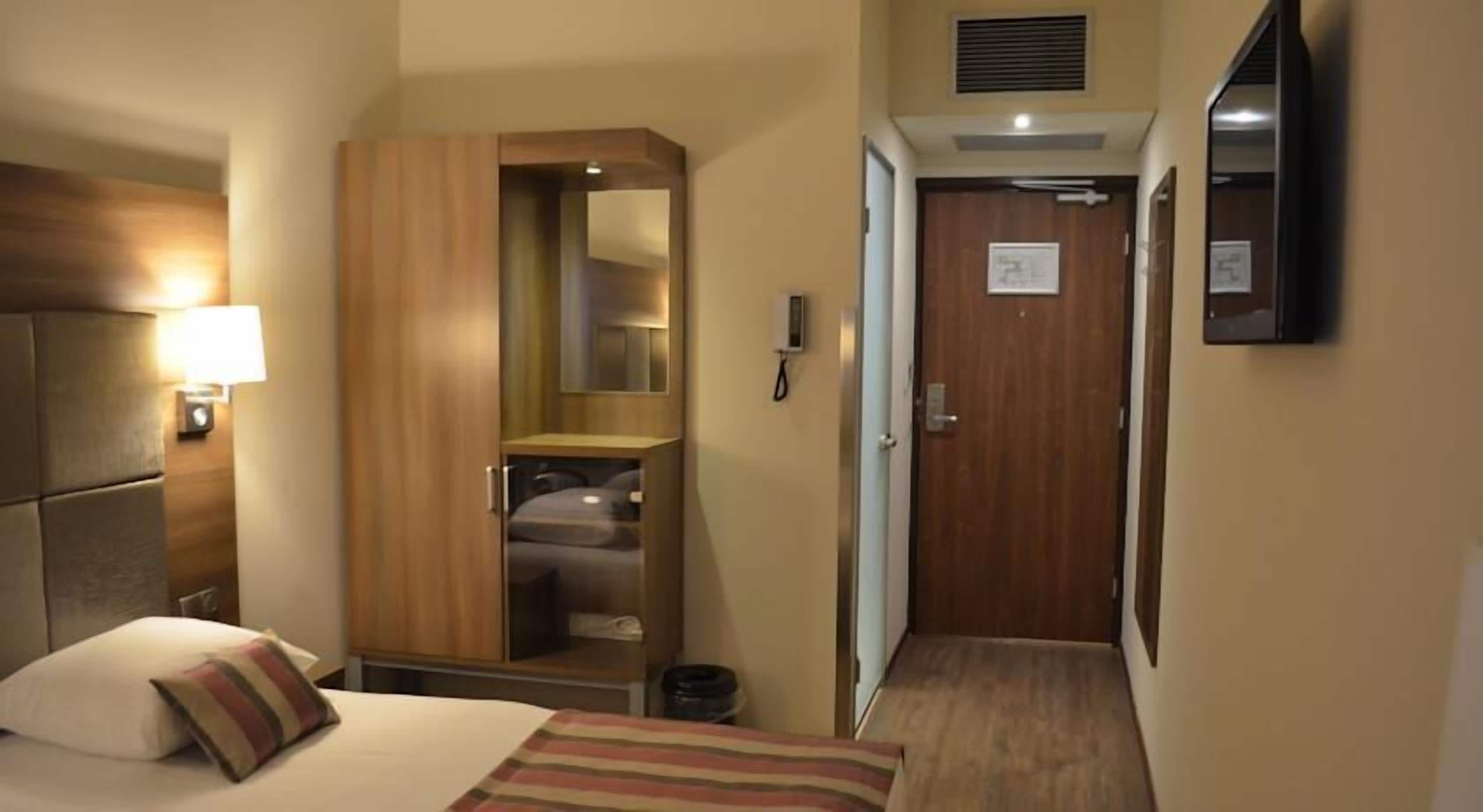 A hotel room with a bed, a wardrobe, a desk, a mirror, a phone, and a door leading to another room.