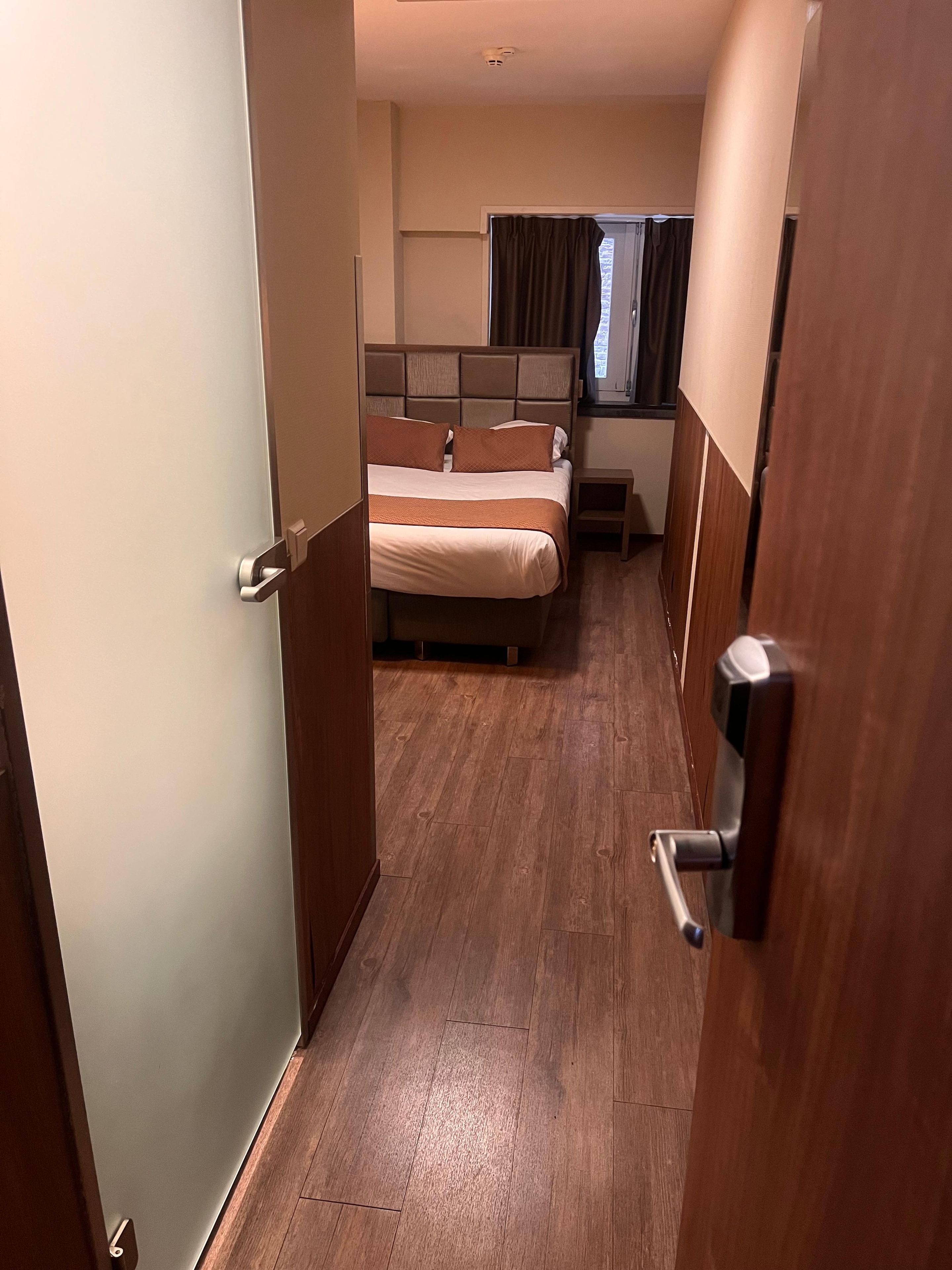 Een smalle gang die leidt naar een slaapkamer met een bed, een nachtkastje en een raam met gordijnen.
