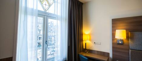 Ein Hotelzimmer mit großem Fenster, einem Schreibtisch mit Lampe und einem Bett mit Kopfteil.