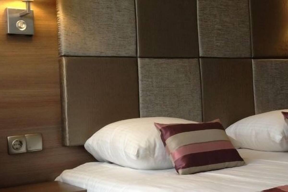 Habitación de hotel con una cama grande, dos almohadas, una mesita de noche, una lámpara de pared y un cabecero de madera.
