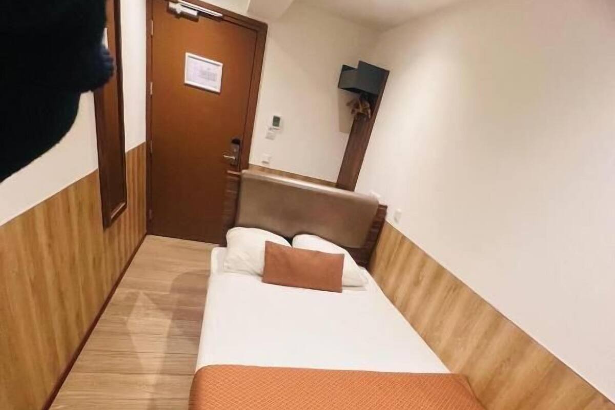 Habitación de hotel con cama, piso de madera, pared marrón, una puerta y televisor de pantalla plana montado en la pared.