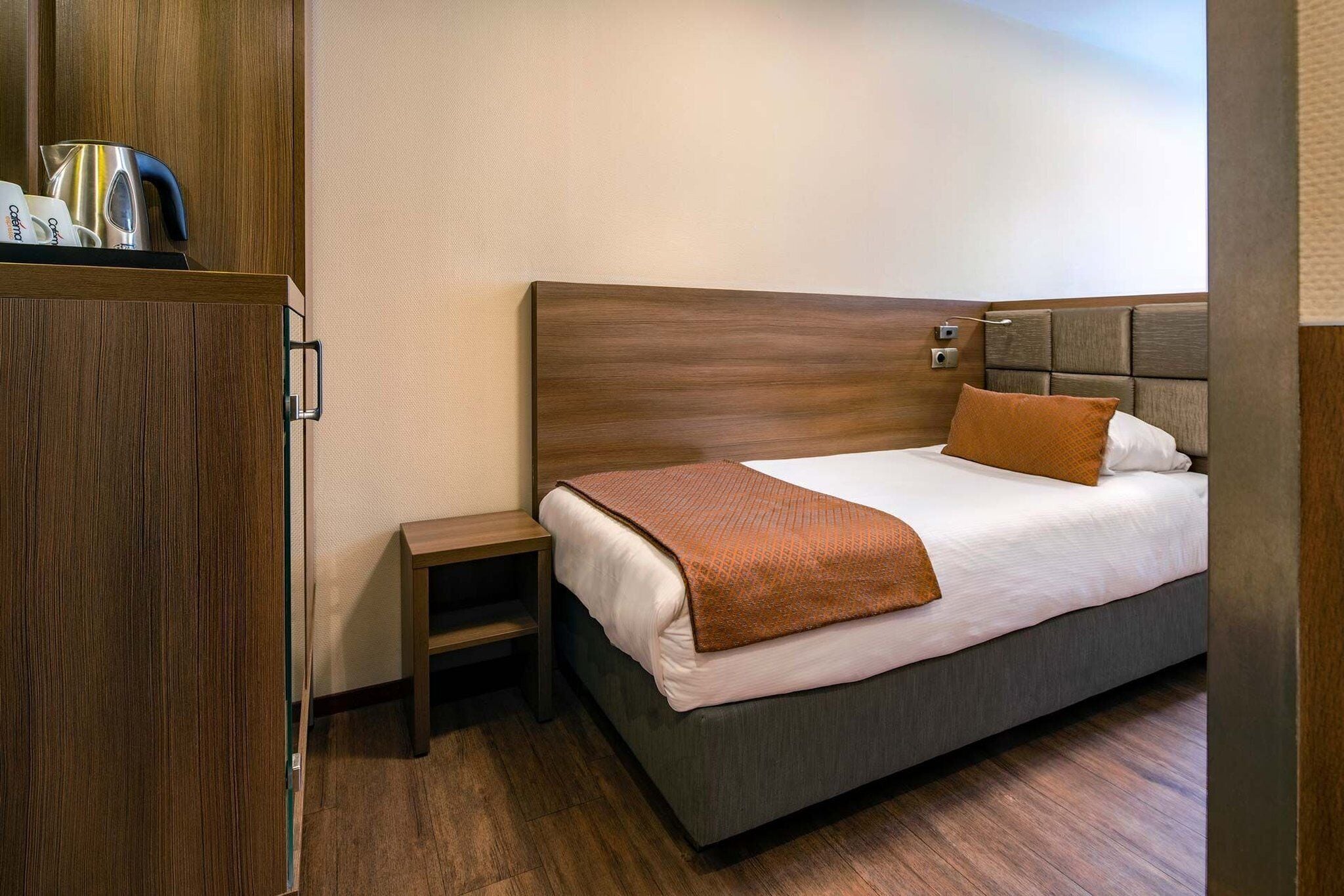 A hotel room with a wooden headboard, a single bed with a white mattress and brown blanket, a small bedside table, and a wooden wardrobe.