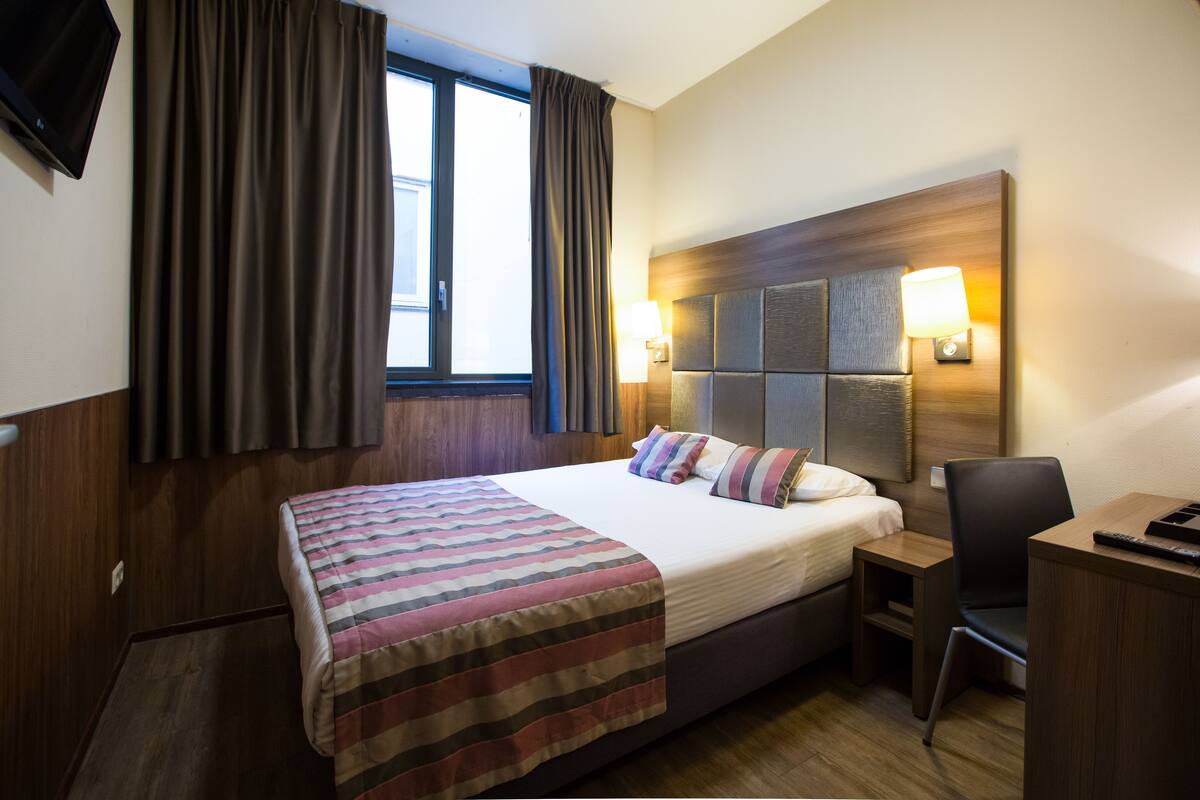 Habitación de hotel con cama, escritorio, silla, televisor y ventana con cortinas.