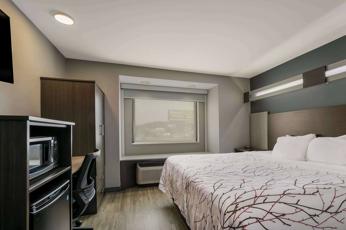 A compact hotel room with a bed, a built-in desk with a microwave, a heater, and a window with a view of buildings.