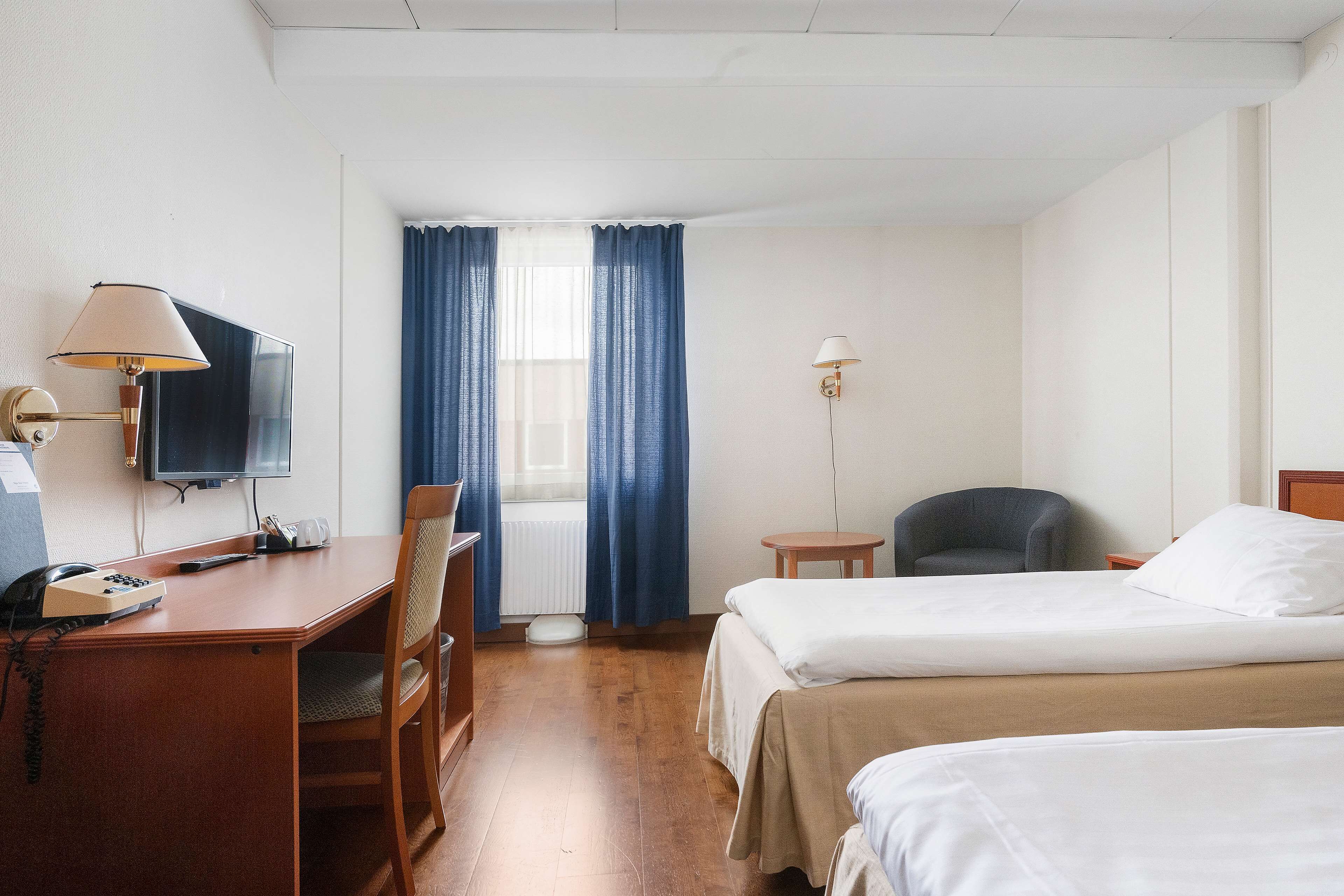 Una camera d'albergo con una scrivania, una sedia, due letti, una TV e una finestra con tende.