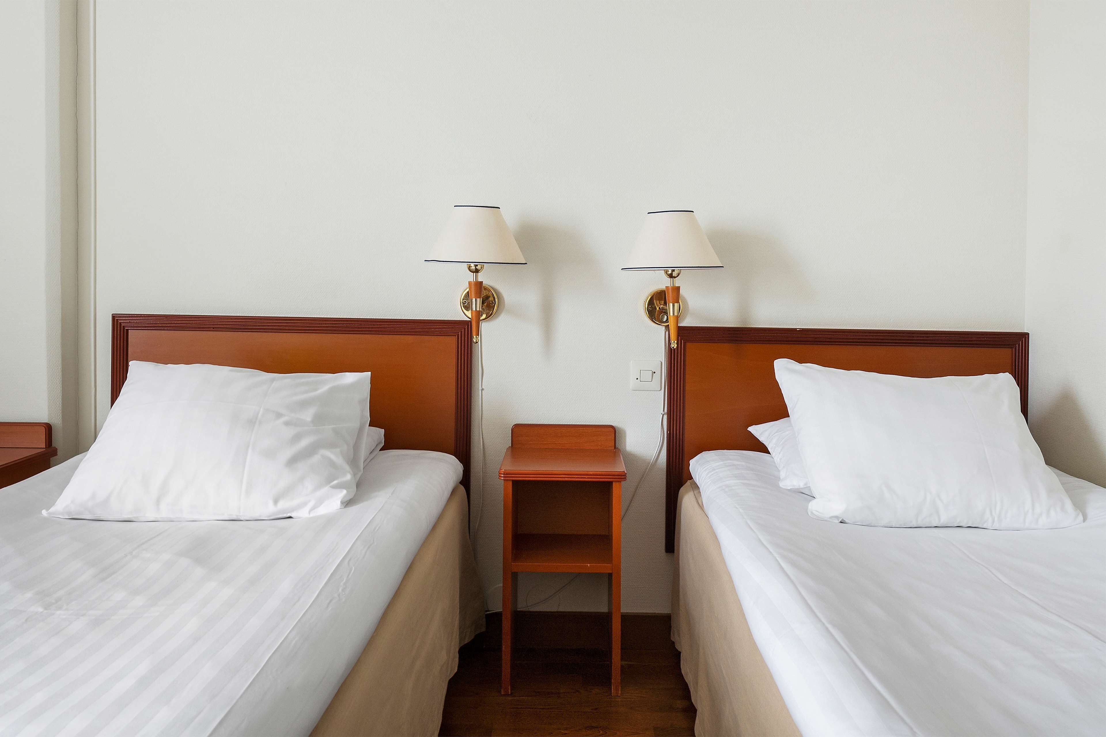 A hotel room with two single beds, each with white linens and wooden headboards. There are two wall-mounted lamps with white shades and wooden bases. A small wooden nightstand is placed between the beds.