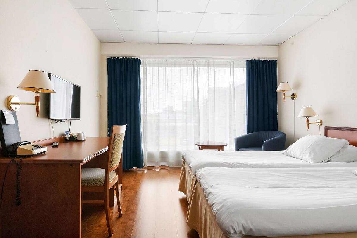 A hotel room with a desk, chair, two beds, a TV, and a window with curtains.