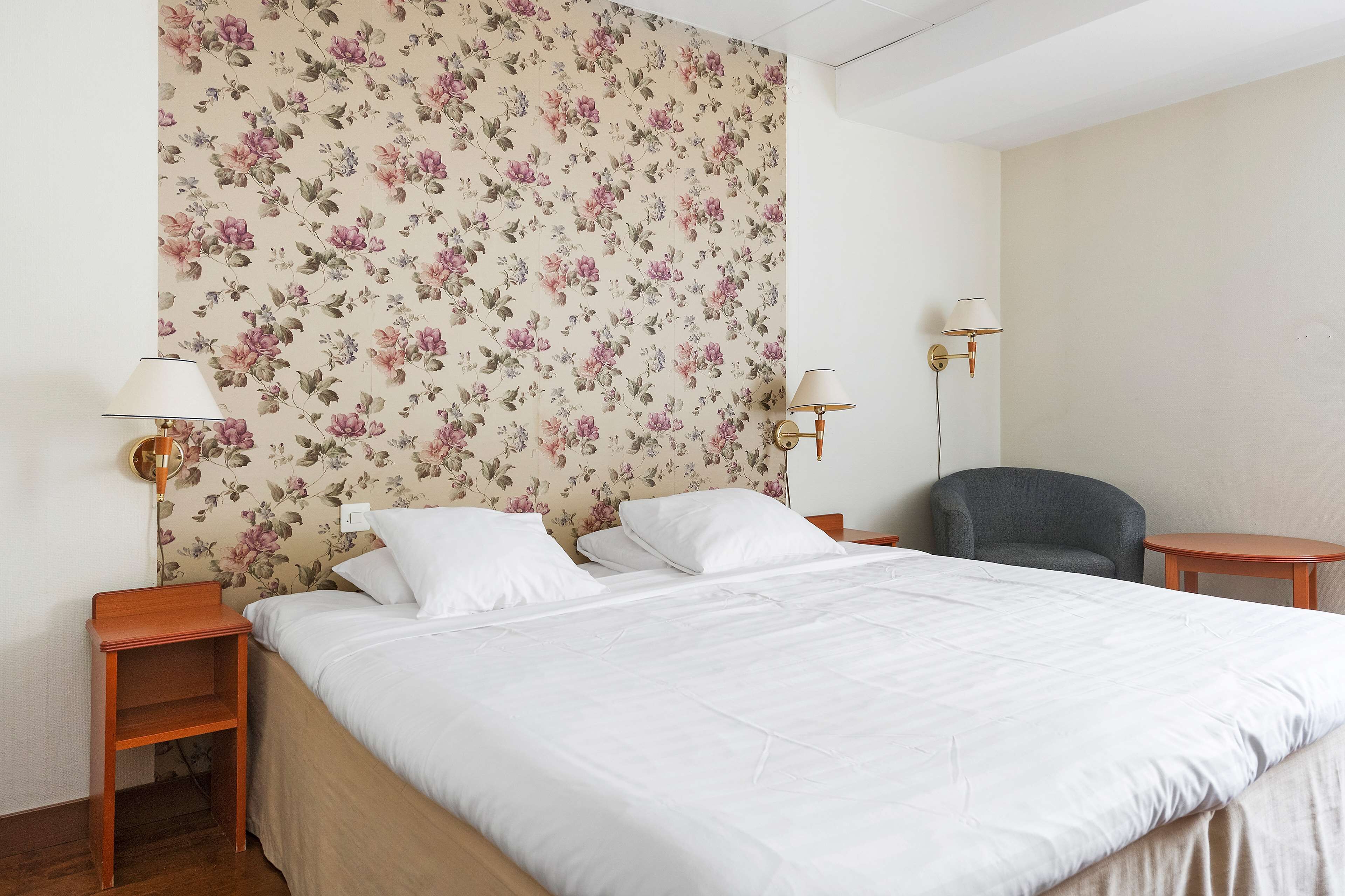 Una cama bien hecha con sábanas blancas, un papel tapiz floral y dos lámparas de noche.