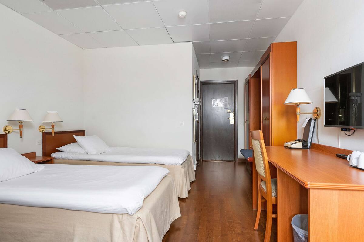 A hotel room with two single beds, a desk with a chair, a television, and a desk lamp.