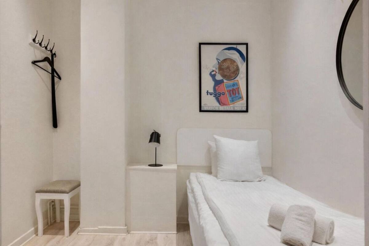 A minimalist bedroom with a bed, a small bedside table, a lamp, a coat rack, and a framed picture on the wall.