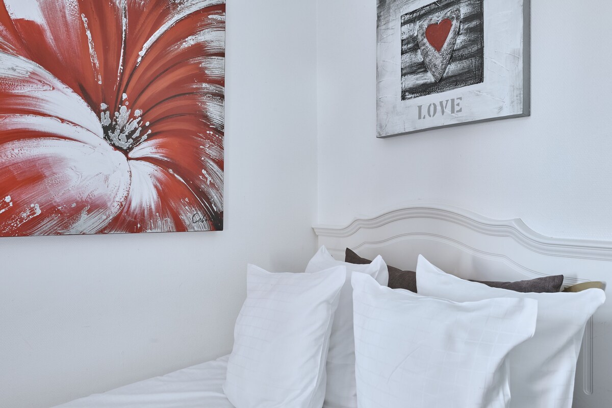 A bed with white bedding, a white headboard, and a red and white painting on the wall.