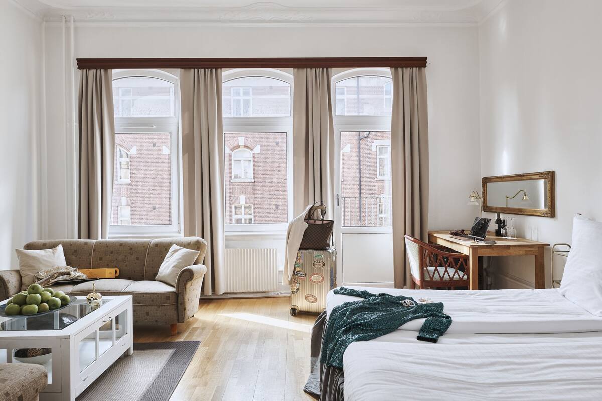 A hotel room with a bed, a desk, a chair, a sofa, and a view of a brick building through large windows.