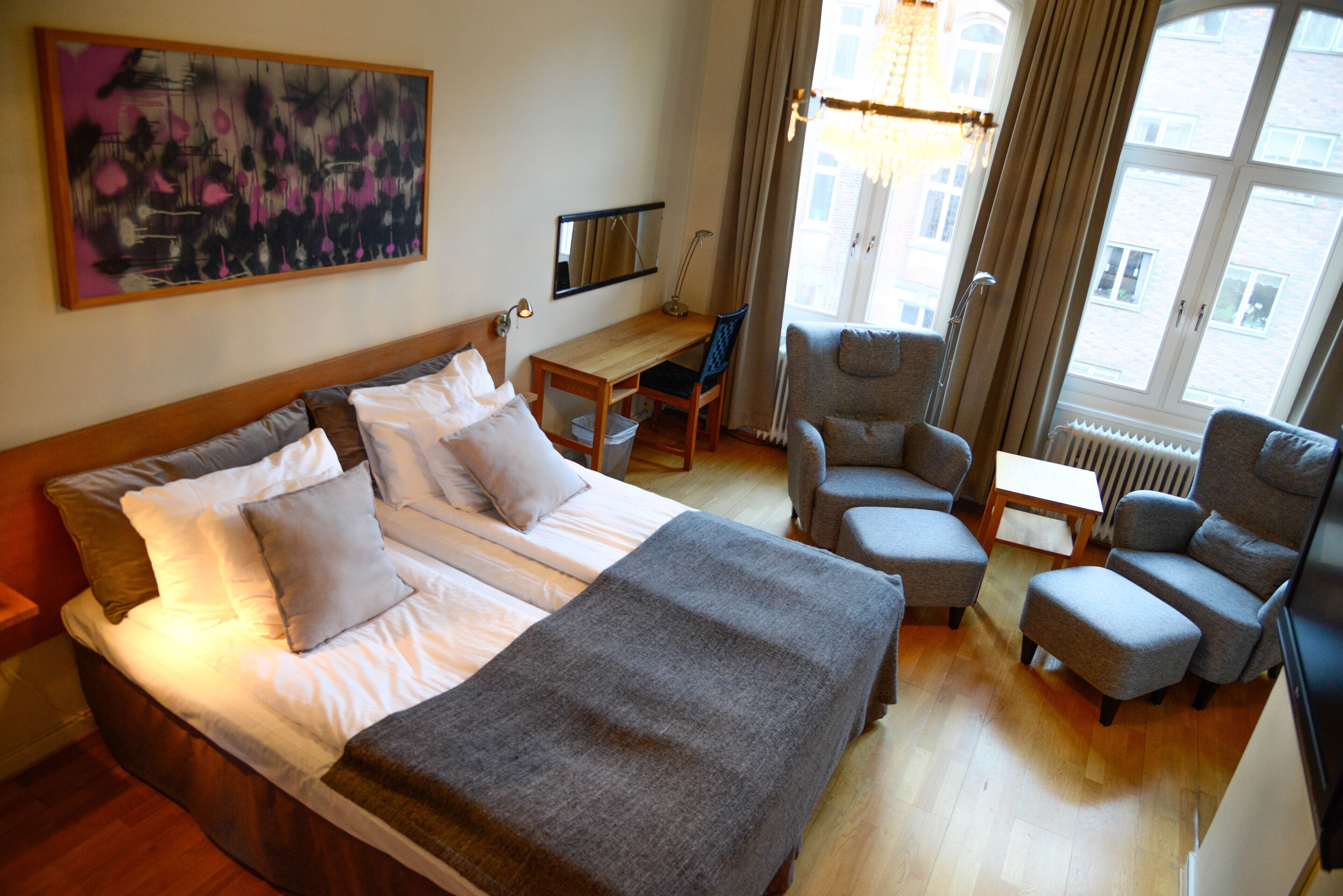 Una habitació d'hotel amb un llit gran, una taula de treball, dues butaques i una finestra amb cortines.