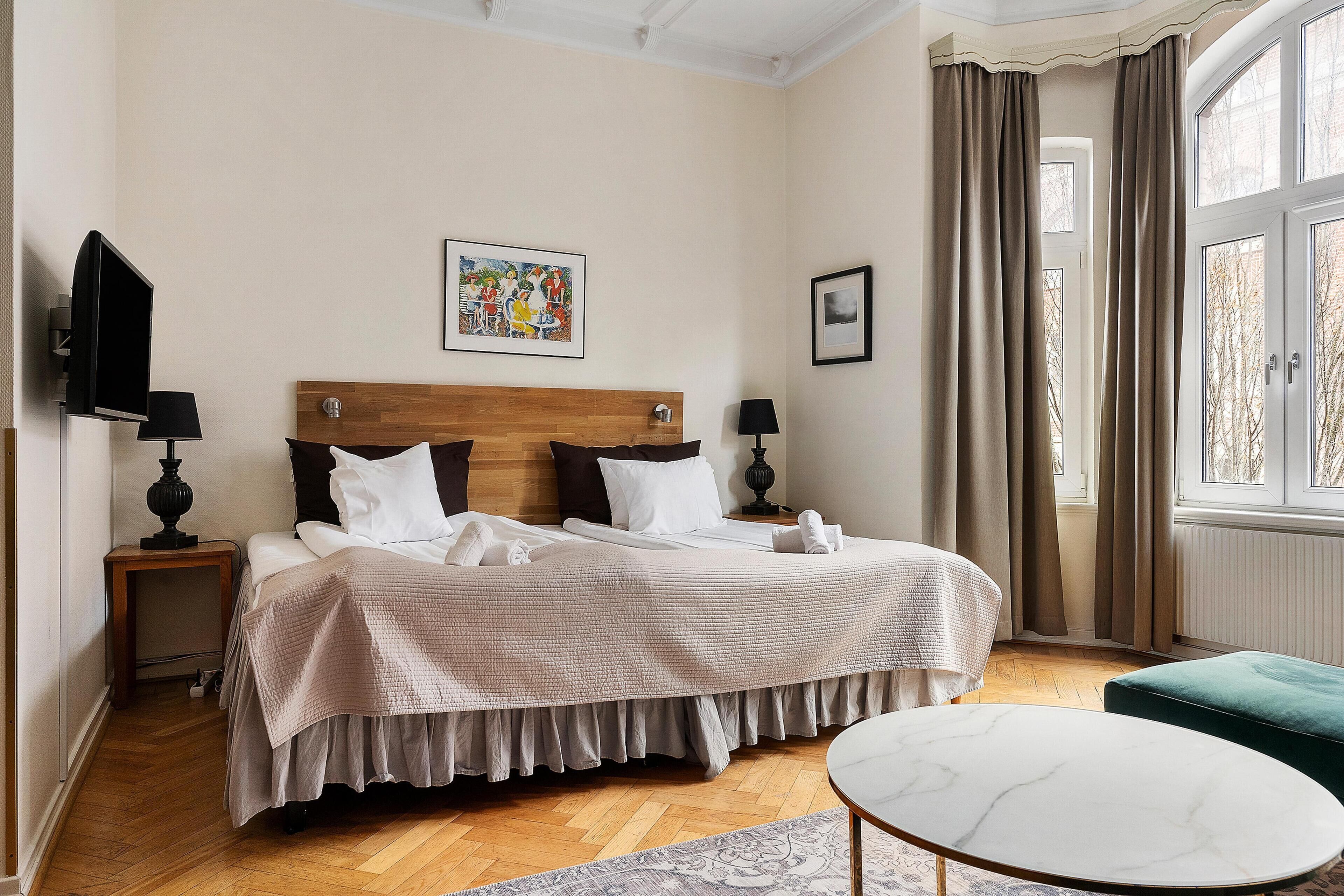 A neatly arranged hotel room with a bed, bedside tables, a TV, a painting, and a window with curtains.