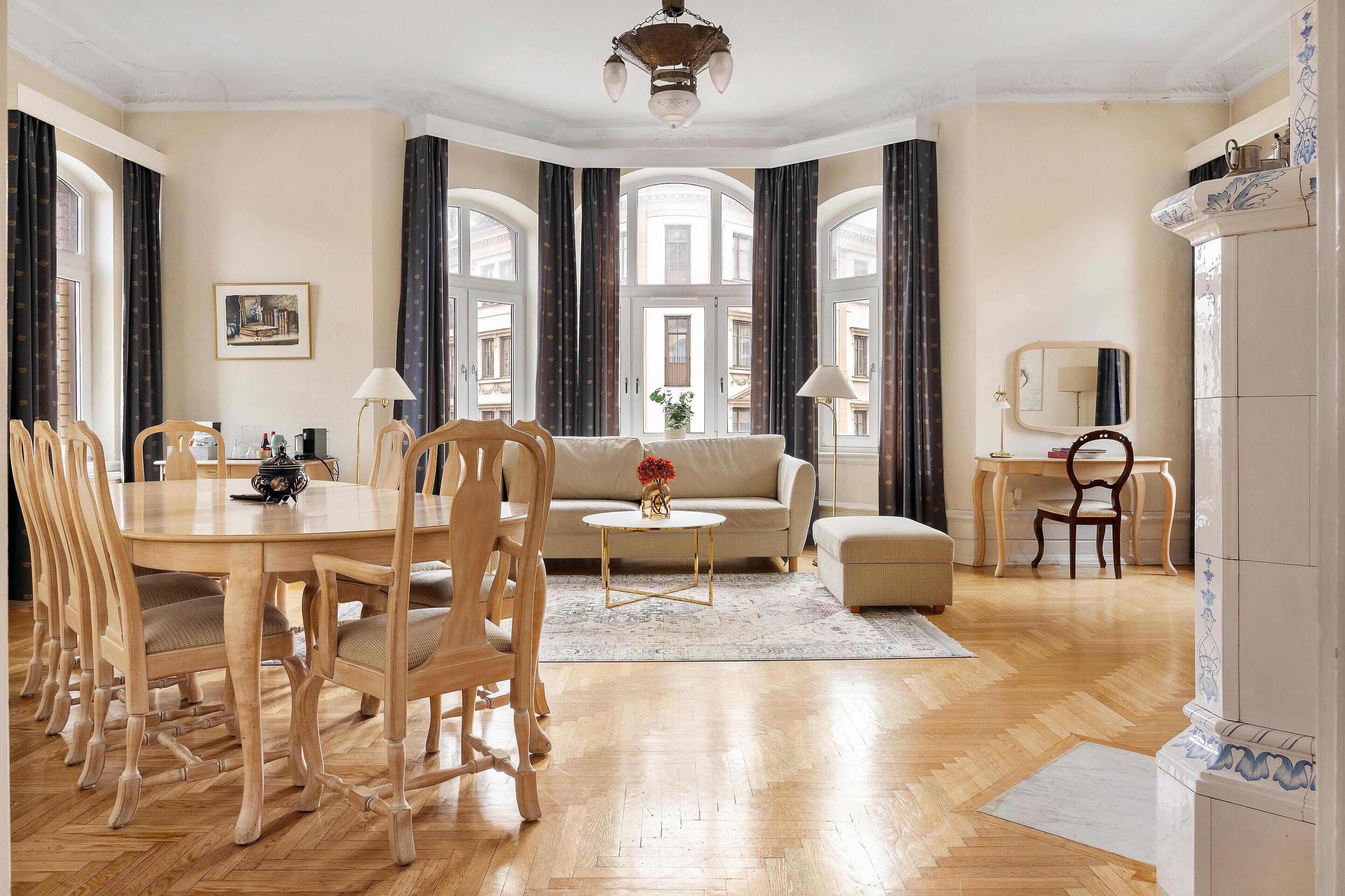 Un amplio salón con mesa de comedor, sofá y sillones, ventanas grandes con cortinas y un candelabro.
