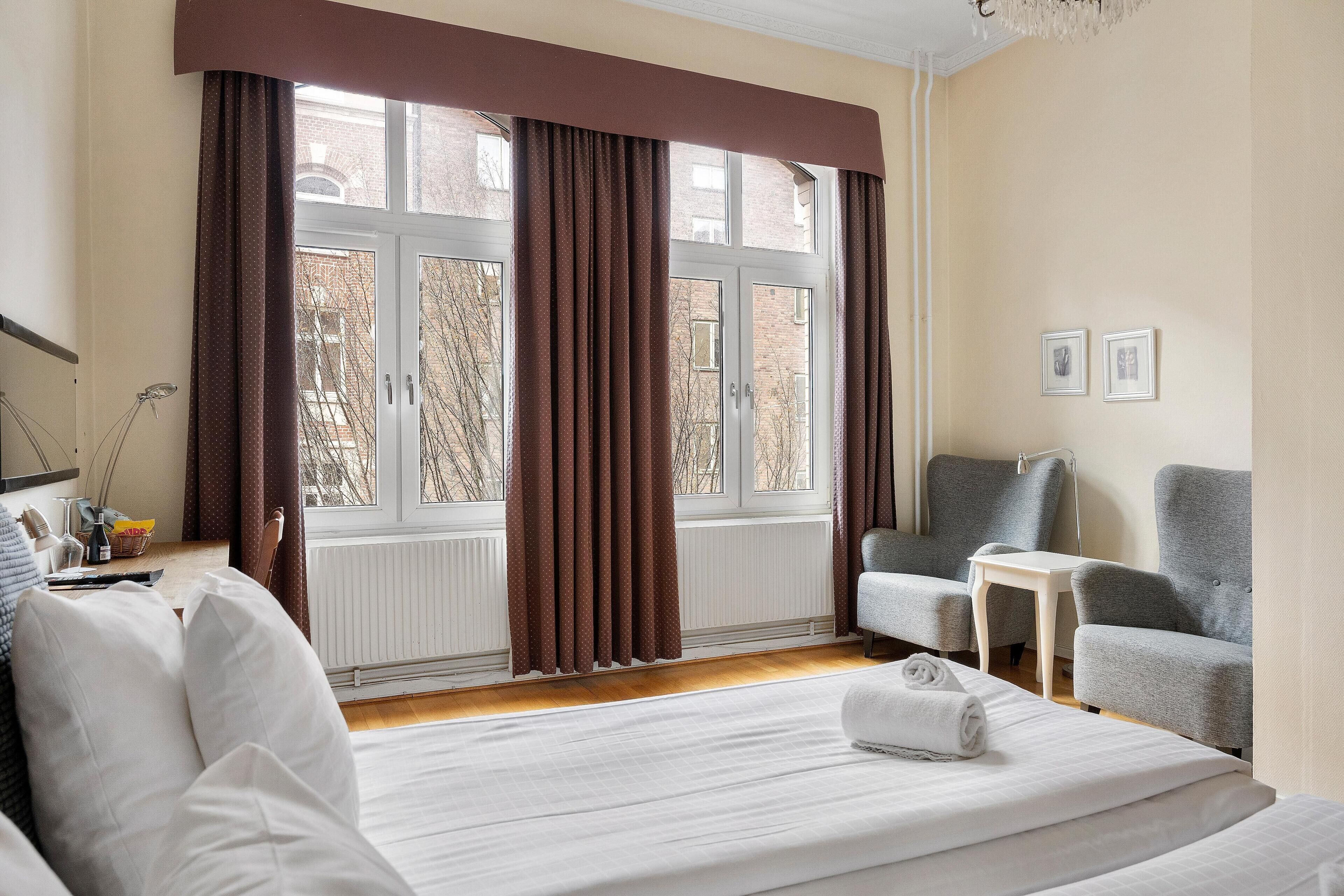 Ein Hotelzimmer mit einem Bett, zwei Sesseln, einem kleinen Tisch und einem Fenster mit Vorhängen.